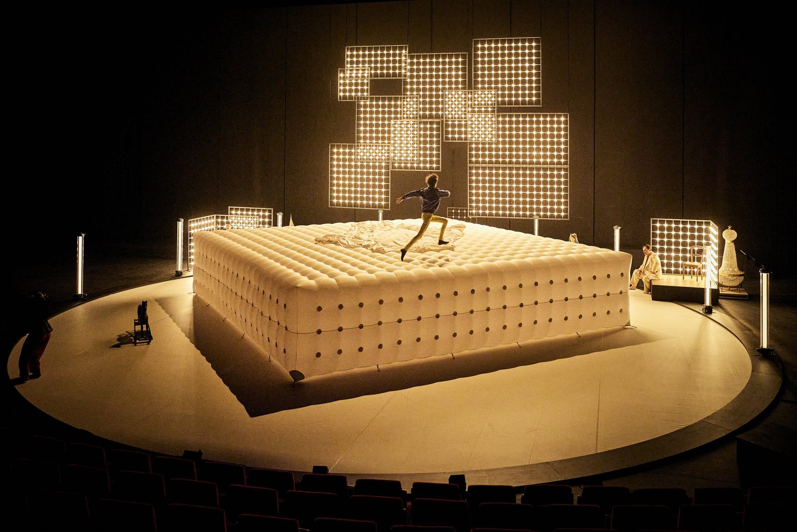 Sur une scène de théâtre, une silhouette bondit sur un immense matelas capitonné et lumineux, devant une sculpture faite de carrés de lumière.