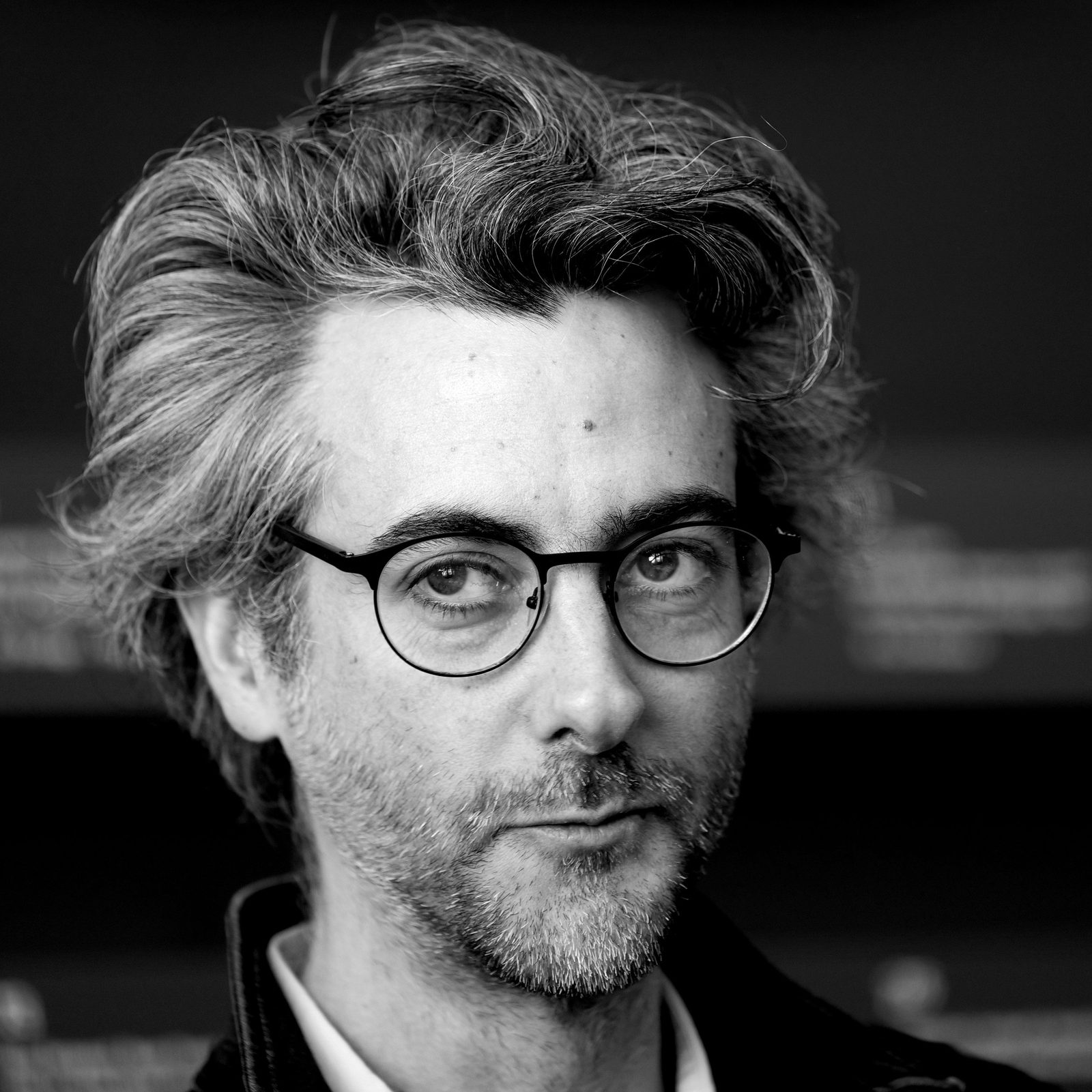 Portrait en noir et blanc d'un homme aux cheveux bouclés et lunettes rondes, regard droit vers l'objectif.