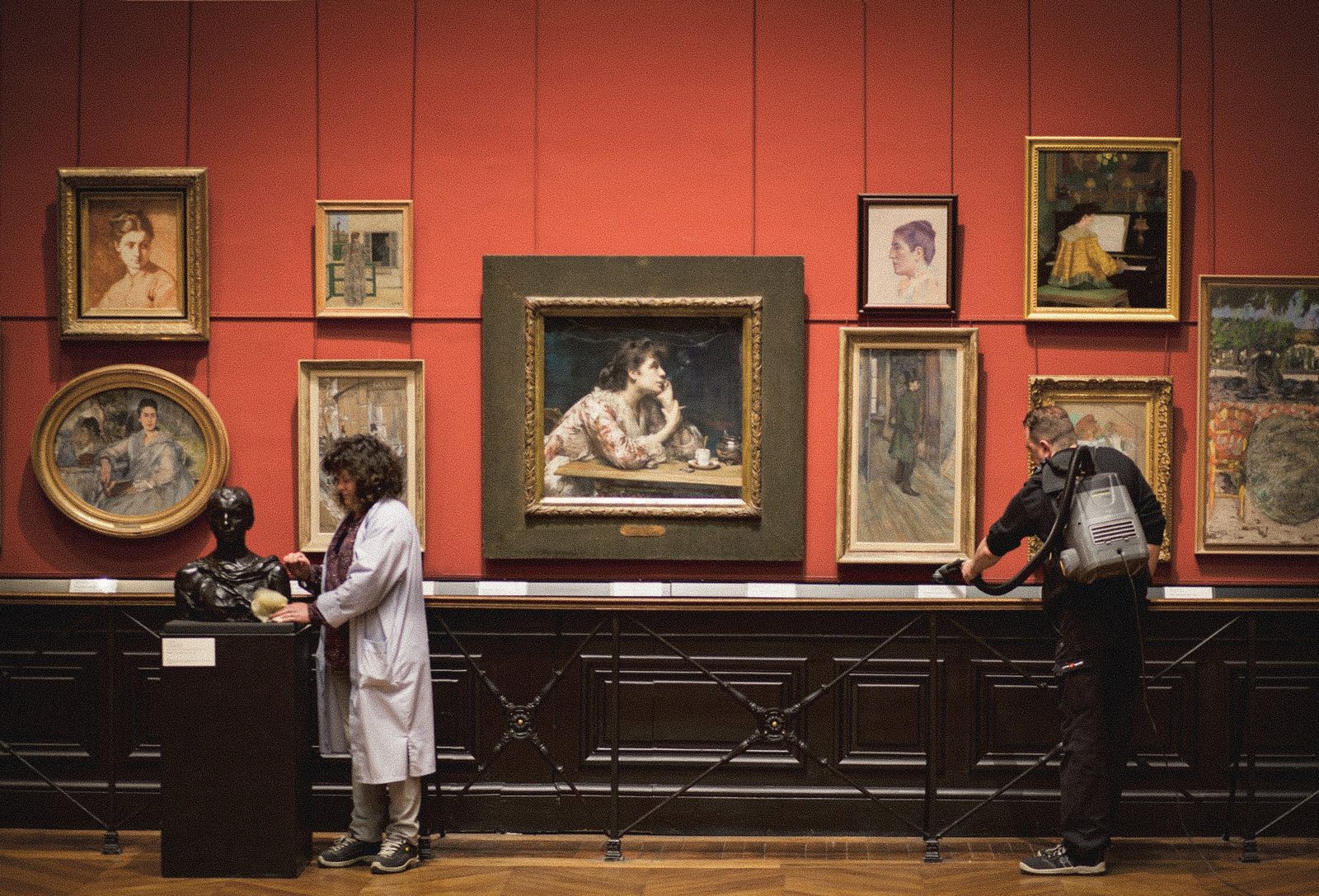 Dans une galerie aux murs rouges couverts de tableaux, deux agents du musée nettoient portraits et sculpture à l'aide d'un pinceau et d'un aspirateur dorsal.