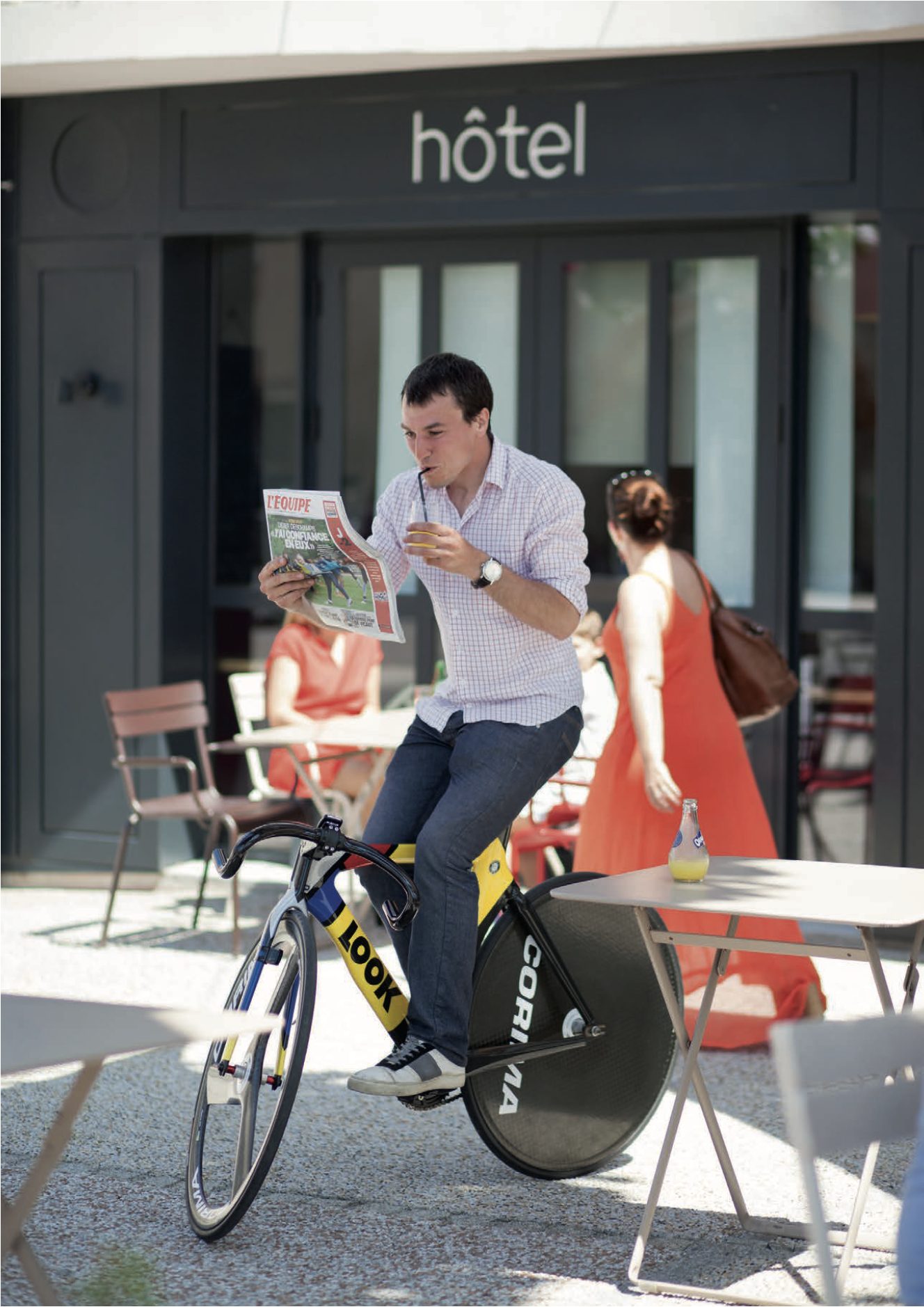 Un homme en équilibre sur un vélo de course lit le journal L'Équipe tout en buvant à la paille, devant un hôtel où une femme en robe orange traverse la terrasse.