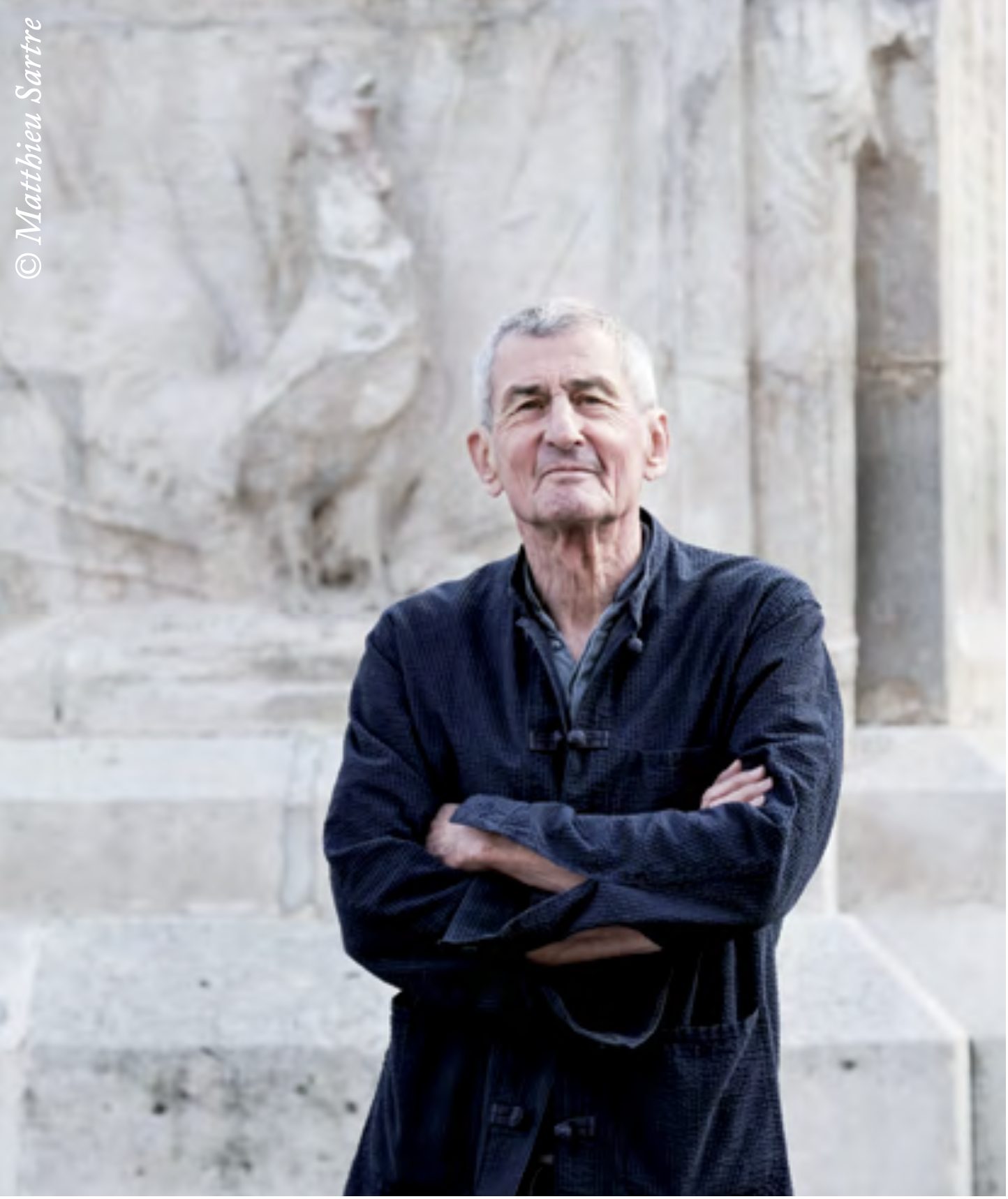Portrait d'un homme aux cheveux gris, bras croisés, en veste sombre, devant les marches et le bas-relief d'un monument de pierre.