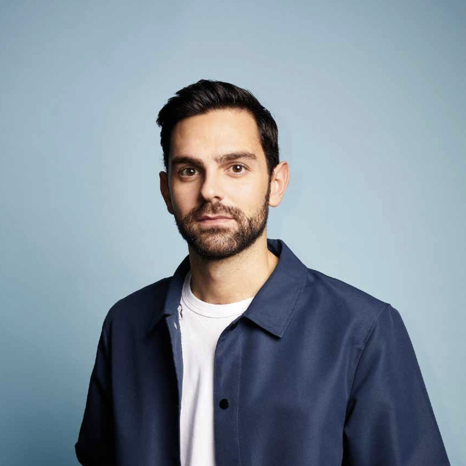 Portrait sur fond bleu : un homme brun à la barbe courte, en veste bleu marine sur un T-shirt blanc.