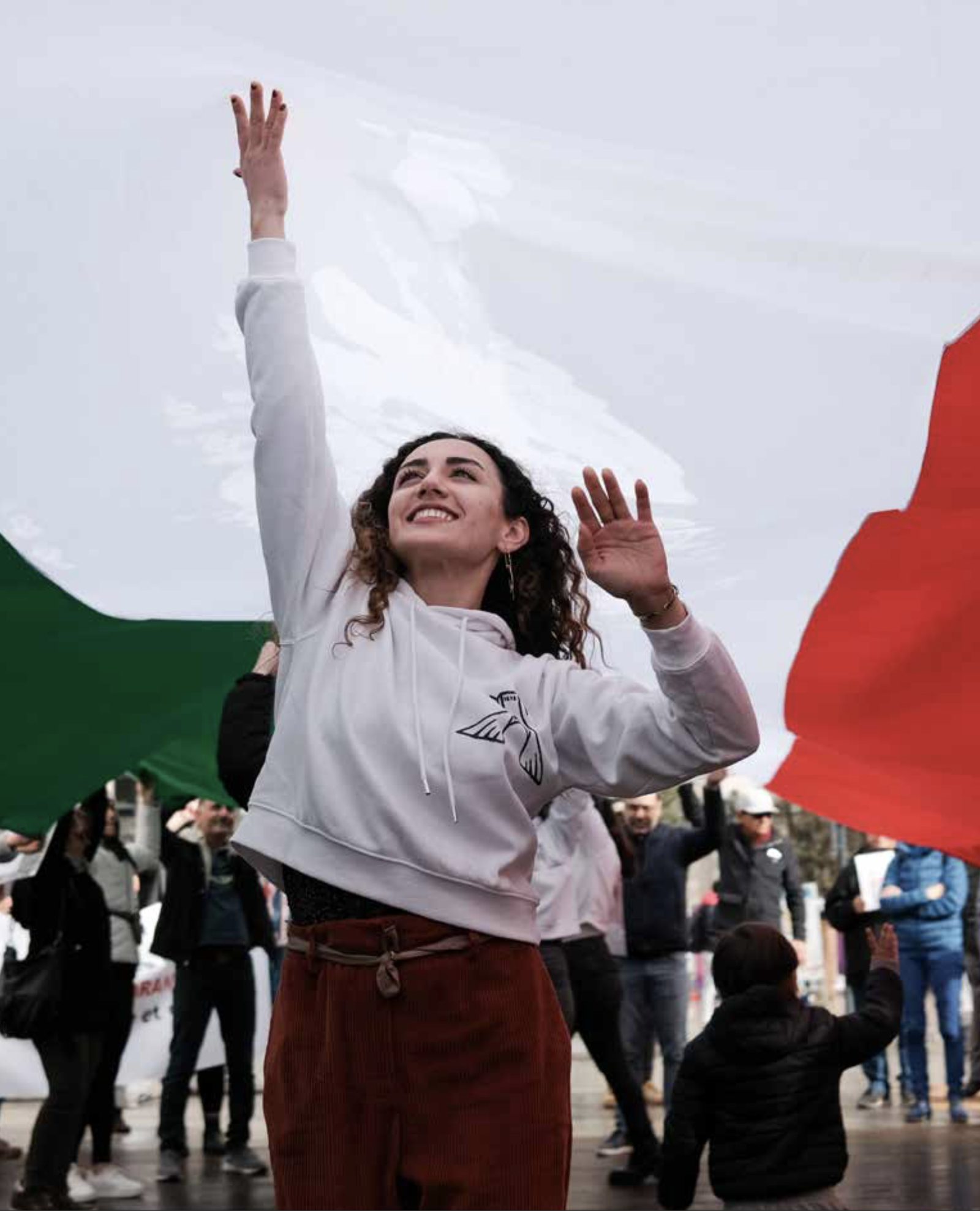 Une jeune femme aux cheveux bouclés, en sweat à capuche blanc, lève les bras en souriant au milieu d'une foule brandissant des drapeaux verts, blancs et rouges.