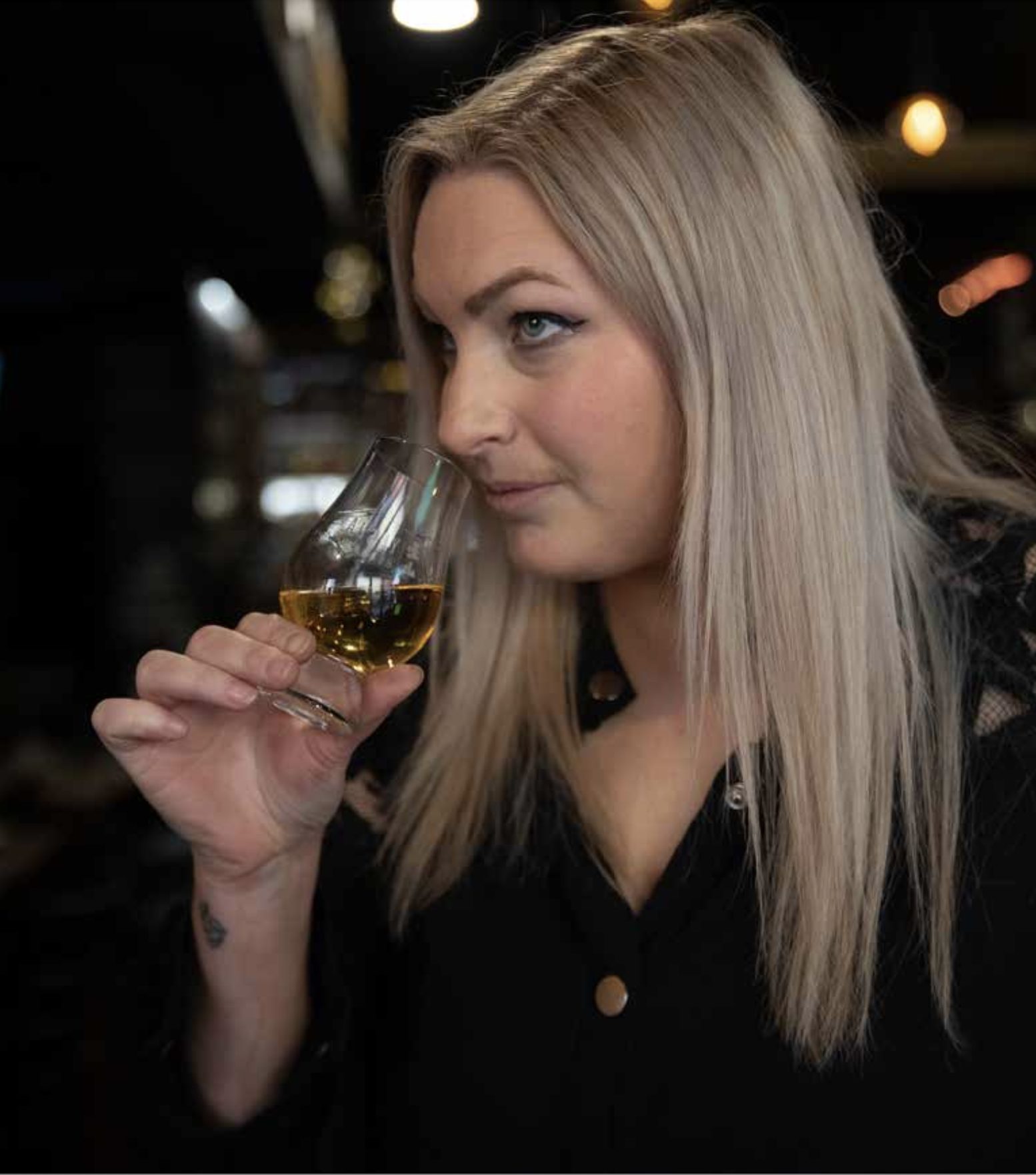 Une femme blonde en haut noir hume un verre de whisky dans un bar à l'éclairage tamisé.