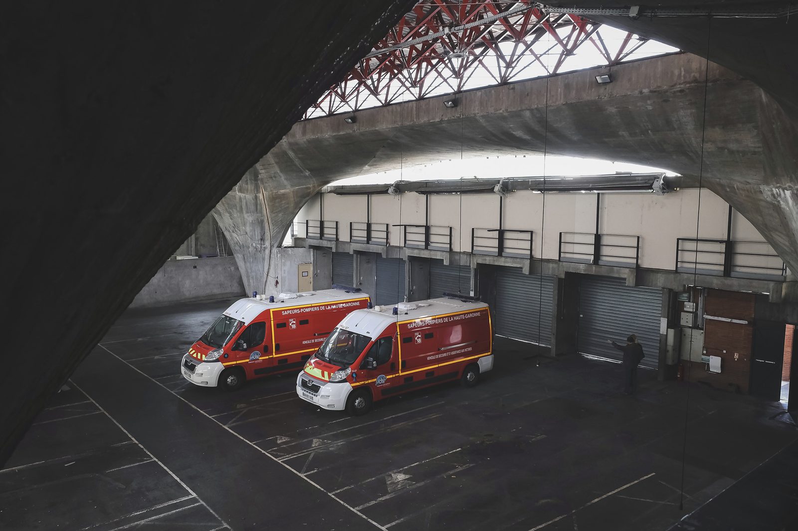 Deux véhicules de secours des sapeurs-pompiers de la Haute-Garonne stationnés dans un vaste hangar aux arches de béton et aux portes métalliques.