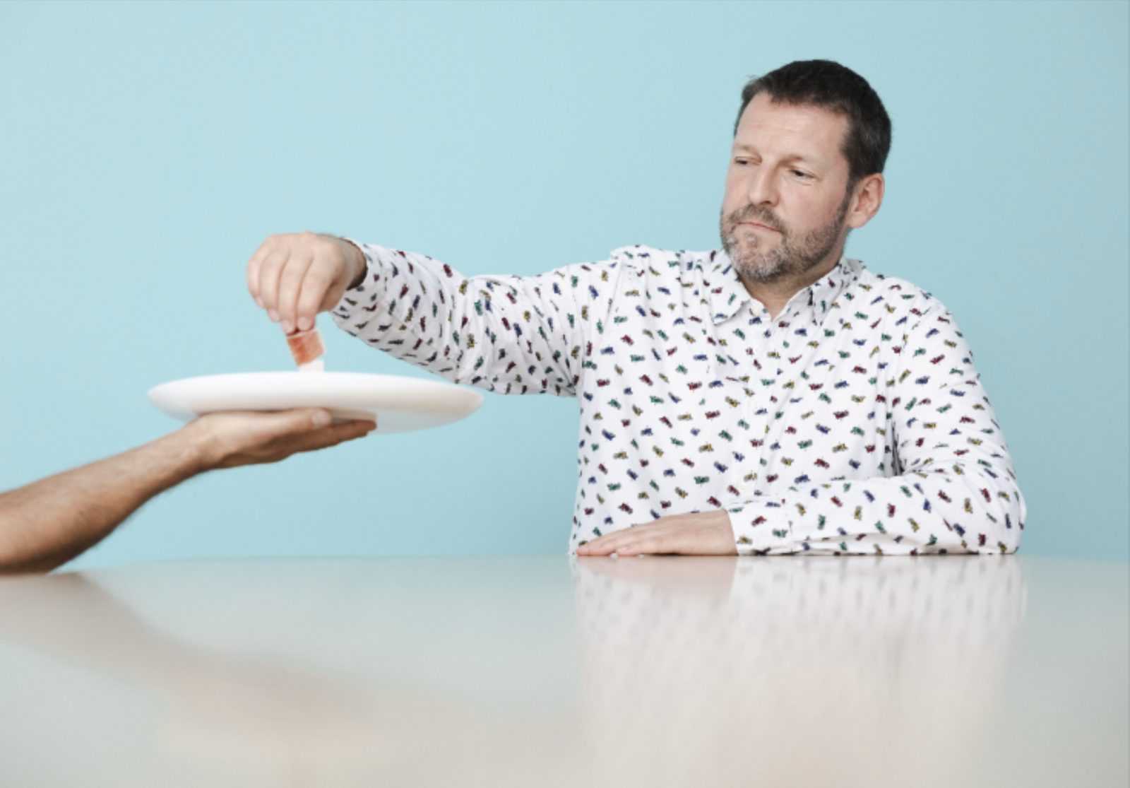 Un homme en chemise imprimée, attablé, s'apprête à goûter un mets présenté sur une assiette tendue par une main, sur fond bleu.