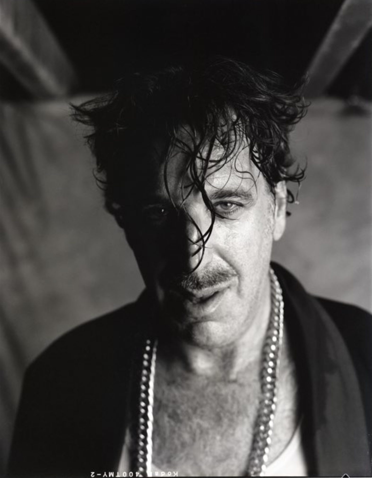 Portrait en noir et blanc à l'éclairage dramatique : un homme aux cheveux bruns en bataille, portant plusieurs colliers, regarde fixement l'objectif.