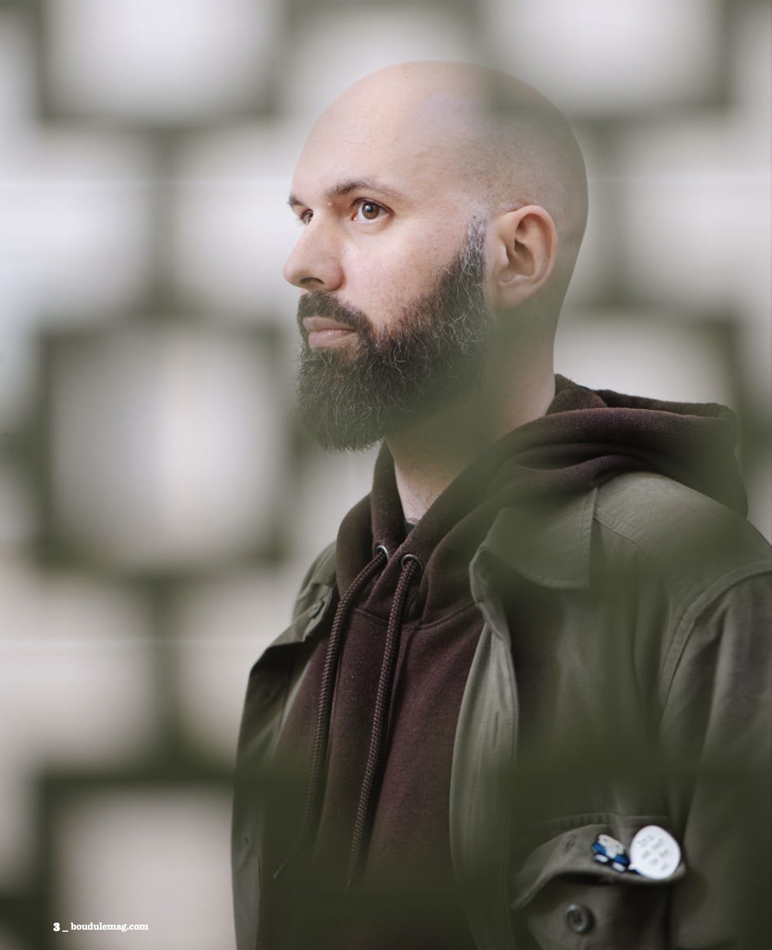 Portrait en double exposition : un homme au crâne rasé et à la barbe brune, en veste kaki, se détache sur un fond de mur à motifs carrés flouté.