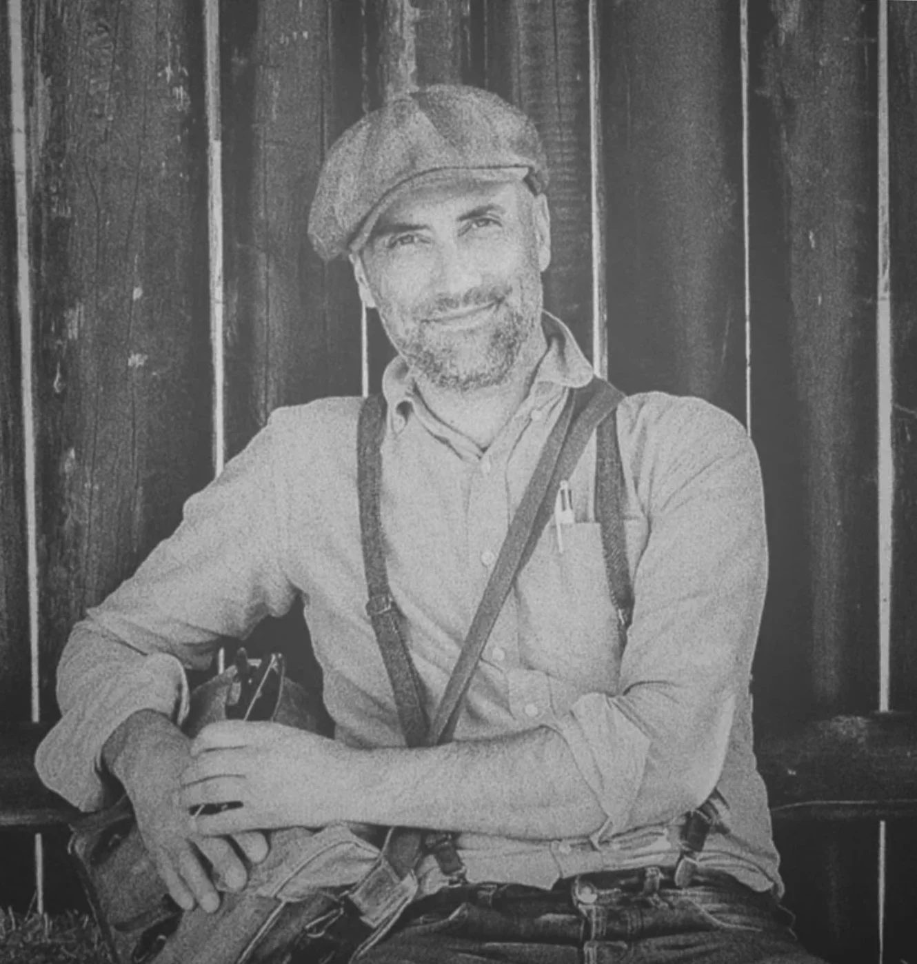 Photo en noir et blanc d'un homme souriant portant une casquette, une chemise, des bretelles et un sac à dos, assis devant un fond en bois.