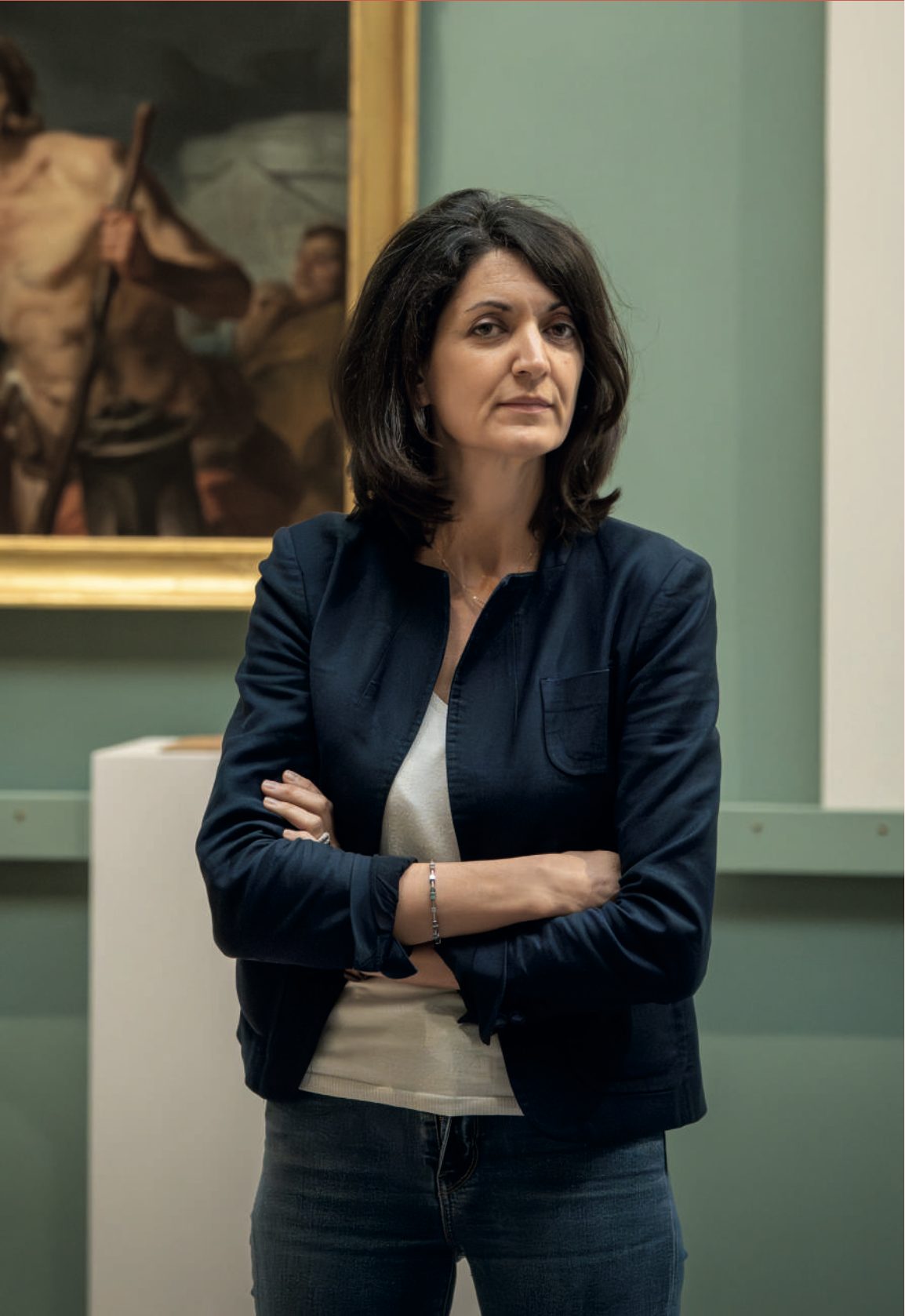 Une femme aux cheveux bruns, veste marine, bras croisés, se tient dans une salle de musée devant un tableau encadré de doré.