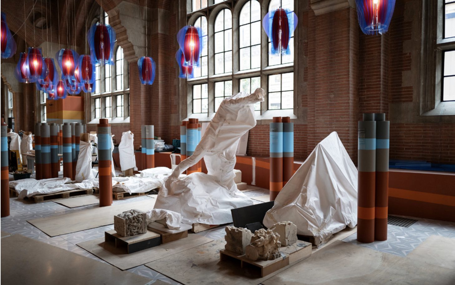 Dans une salle voûtée en briques, des sculptures emballées de housses blanches sont exposées sous des suspensions de verre bleu et rouge.