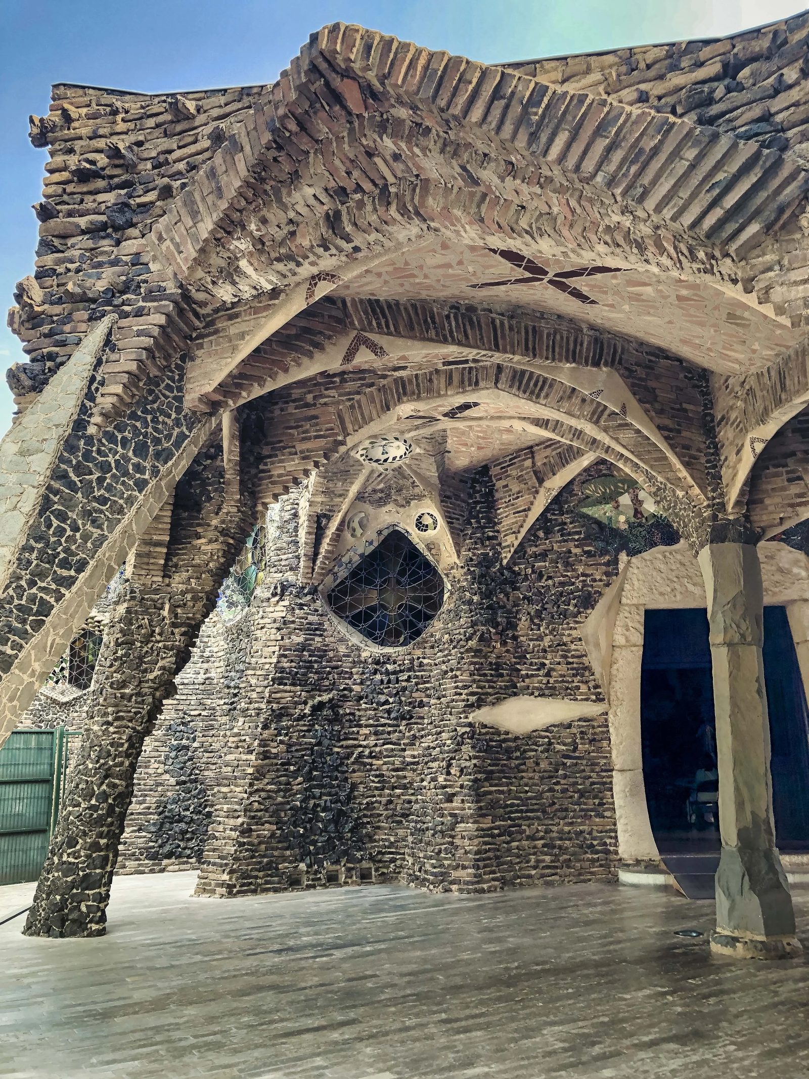 Vue architecturale des voûtes de pierre et de brique aux formes organiques, typiques du style de Gaudí, avec vitraux et colonnes inclinées.