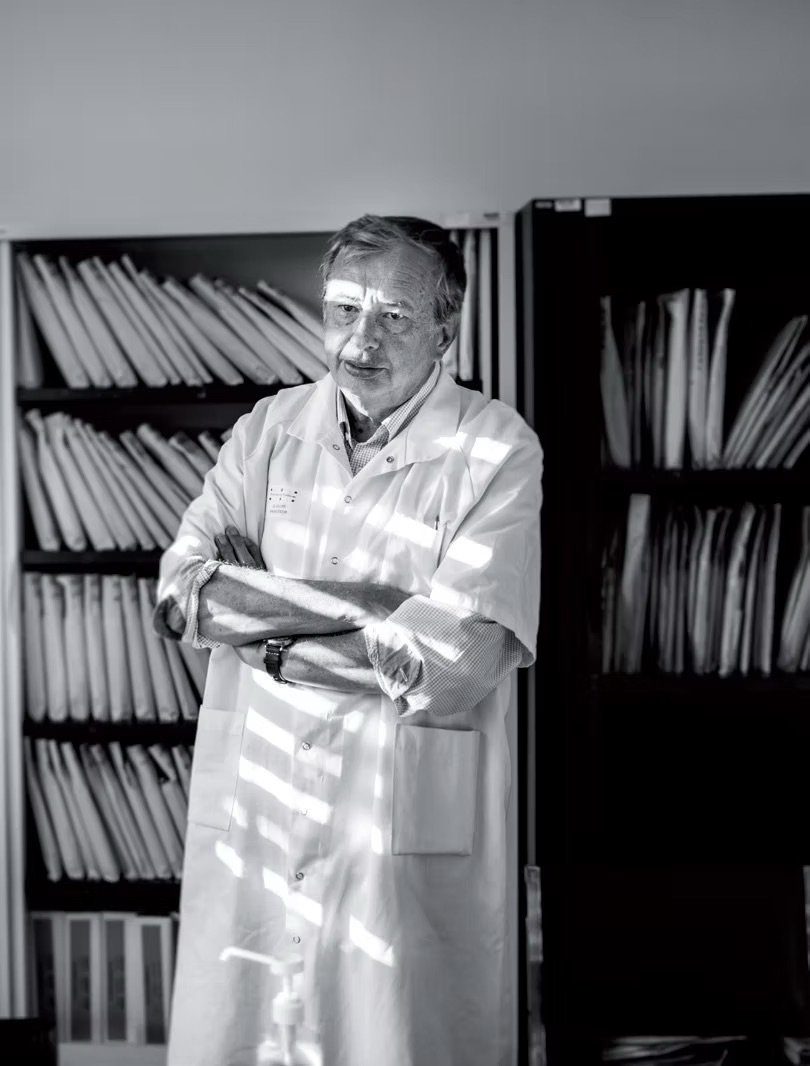 Portrait en noir et blanc : un homme âgé en blouse blanche, bras croisés, se tient devant des étagères d'archives et de documents roulés.