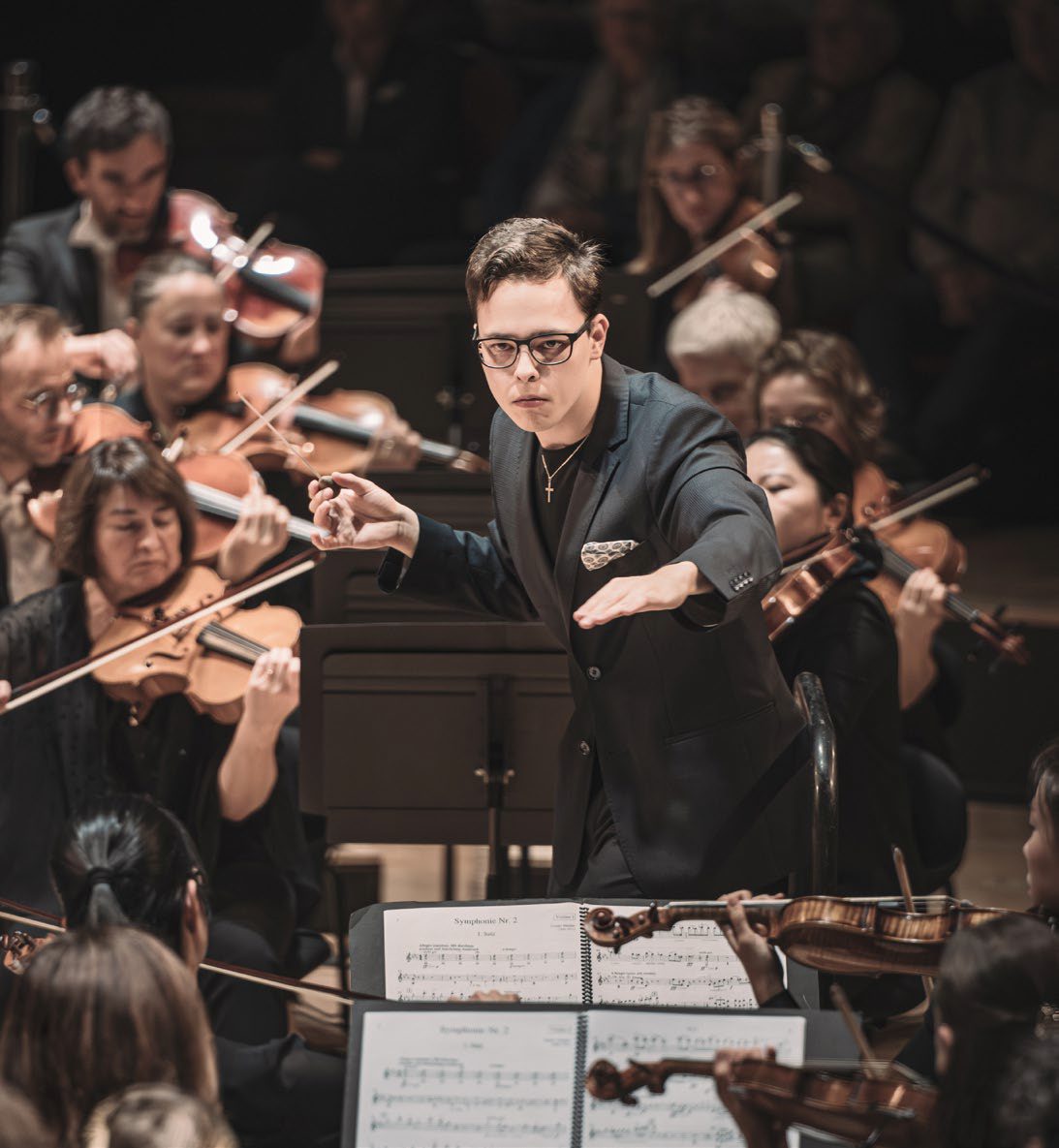 Un jeune chef d'orchestre à lunettes, en costume sombre, dirige avec sa baguette au milieu de musiciens jouant du violon.