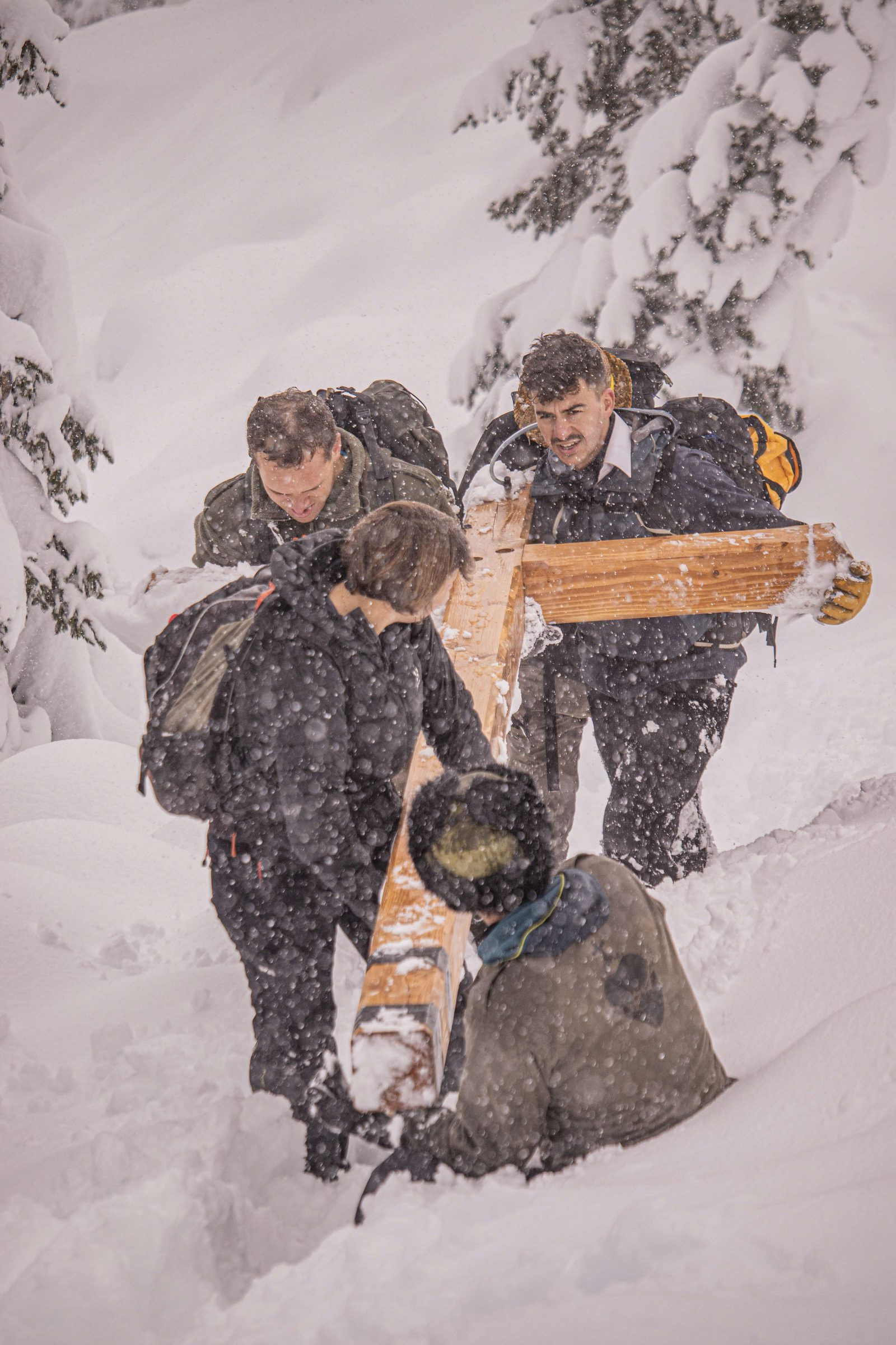 Quatre porteurs hissent une croix de bois dans la neige, sous une chute de flocons, au milieu des sapins enneigés.