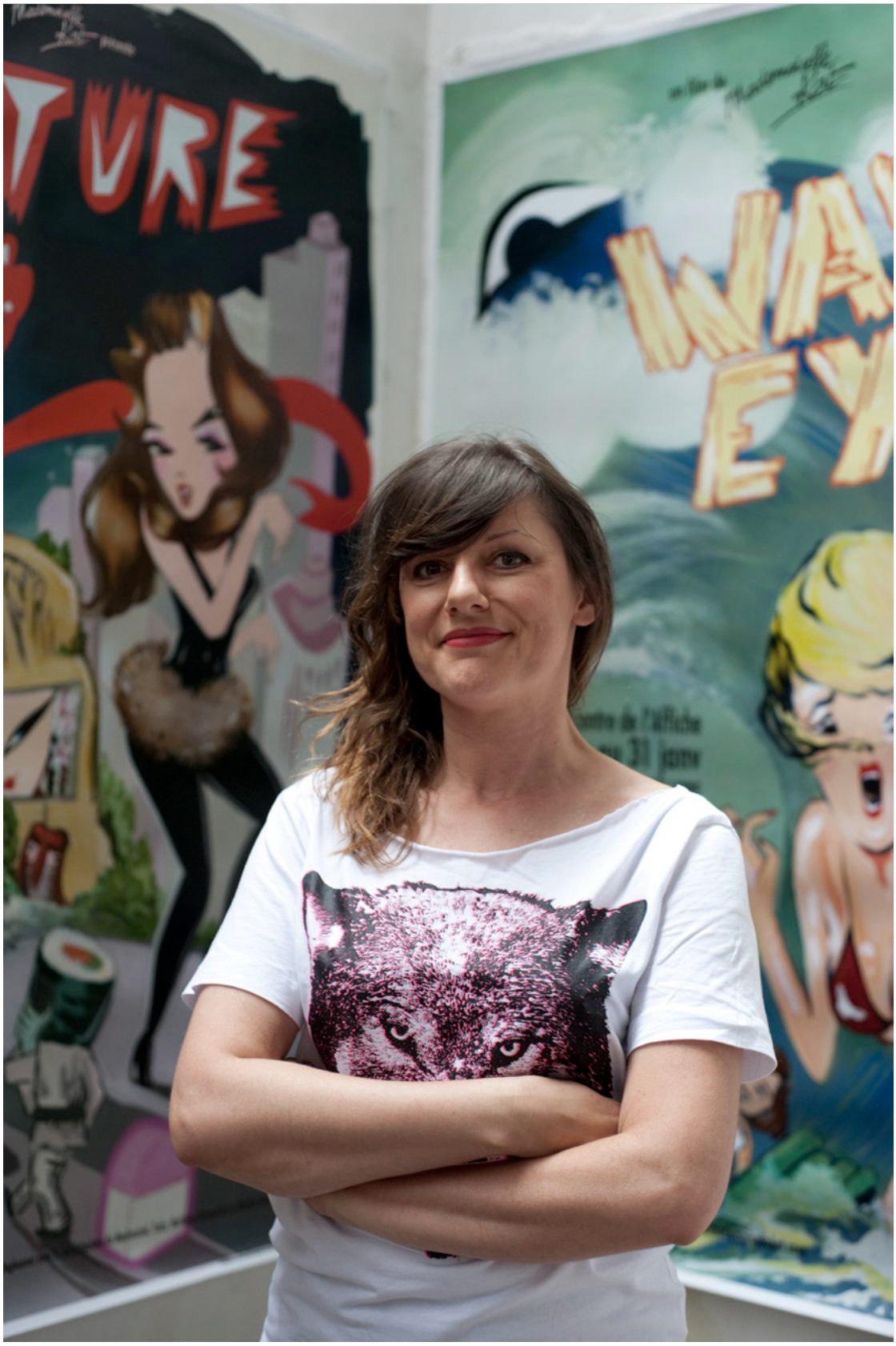 Portrait de l'artiste Mademoiselle Kat, bras croisés, tee-shirt blanc à motif de loup, devant deux affiches peintes de son atelier.