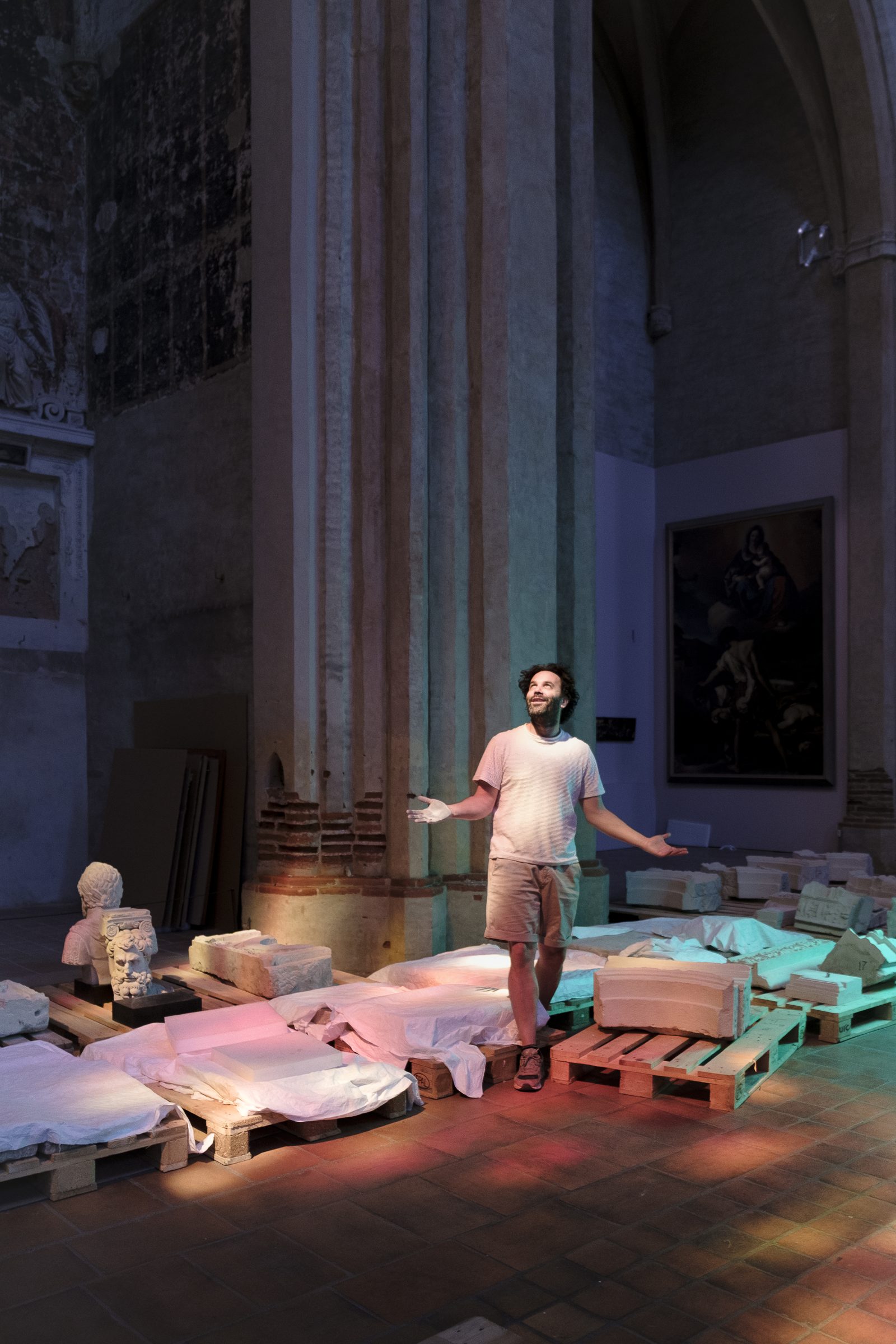 Nicolas Dhervillers, bras ouverts, debout au milieu des plâtres du musée des Augustins entreposés sur palettes, sous des éclairages colorés, dans la nef de l'église.