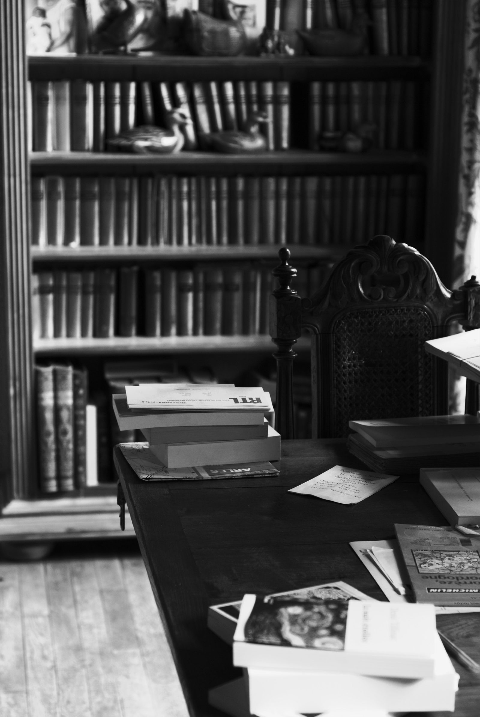 Le bureau de Denis Tillinac à Auriac : une table de bois sombre couverte de livres, de cartes routières et de papiers, devant une chaise sculptée et une bibliothèque de volumes reliés.