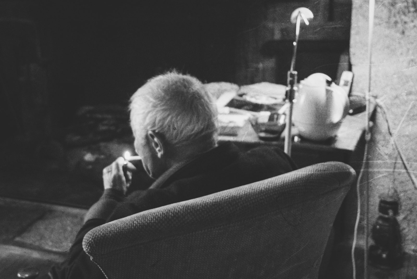 Denis Tillinac, de dos dans un fauteuil, allume une cigarette dans la pénombre de sa maison d'Auriac, face à une table encombrée éclairée par une lampe.