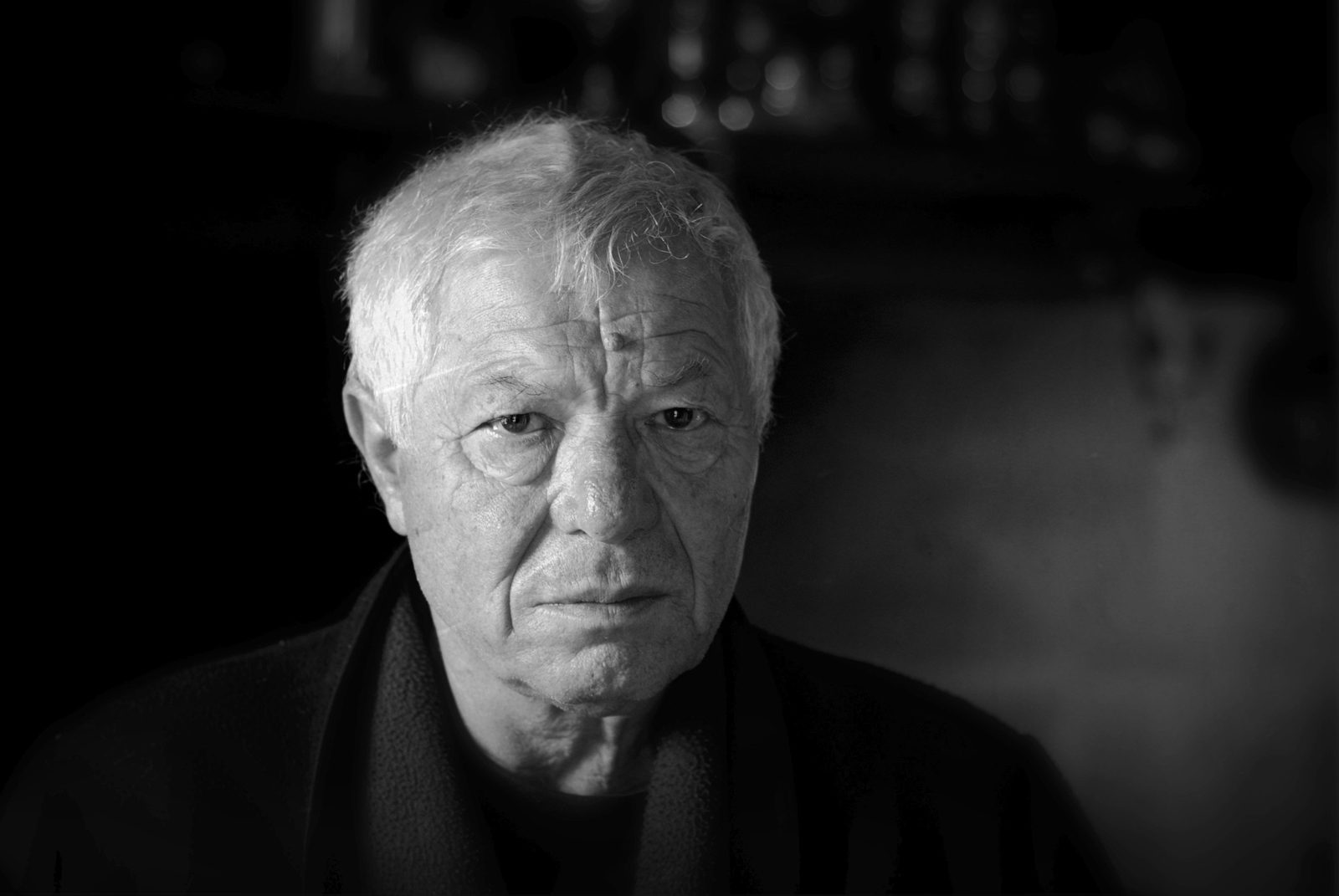 Portrait en noir et blanc de Denis Tillinac, cheveux blancs en bataille, regard grave tourné vers l'objectif, sur un fond sombre.
